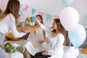 Trois femmes assises partagent des boissons dans des tasses colorées lors d'une baby shower, entourées de ballons et de guirlandes.